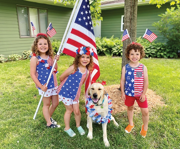 Kids decked out in patriotic clothing pose with the American flag and a dog.