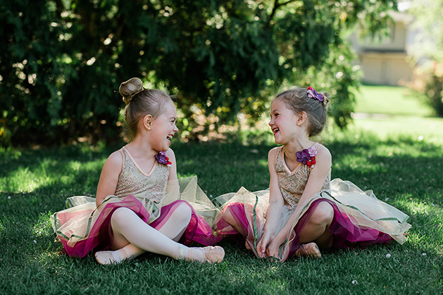 Two young ballerinas sit in the grass, laughing.